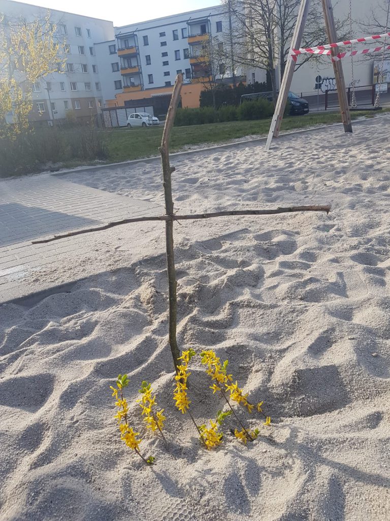 Holzkreuz aus Ästen im Sand steckend mit Forsythie